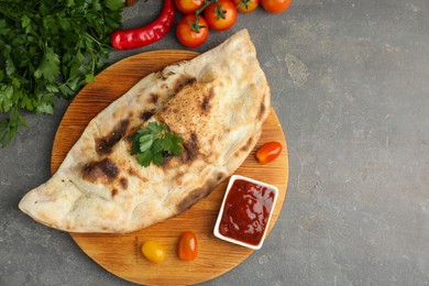 Photo of Board with tasty calzone, tomatoes, sauce and parsley on grey textured table, flat lay. Space for text