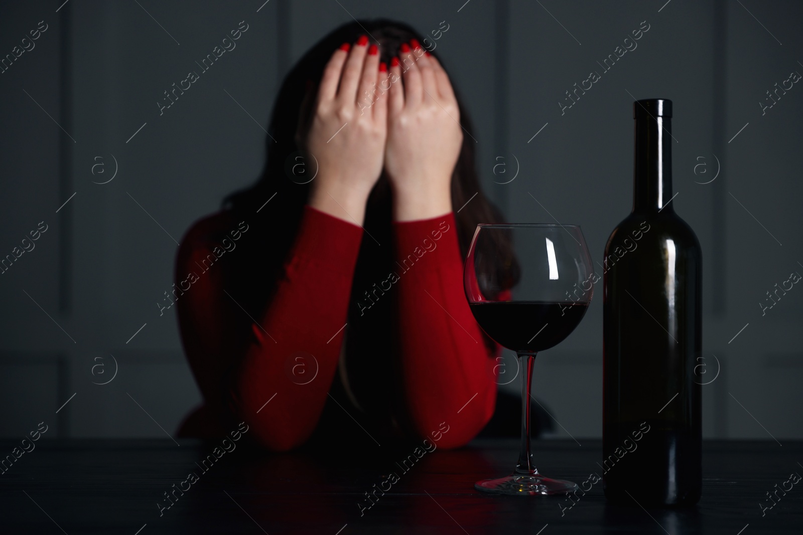 Photo of Alcohol addiction. Woman covering her face at wooden table indoors, focus on glass of red wine and bottle