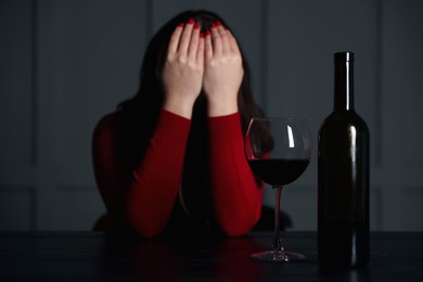 Photo of Alcohol addiction. Woman covering her face at wooden table indoors, focus on glass of red wine and bottle