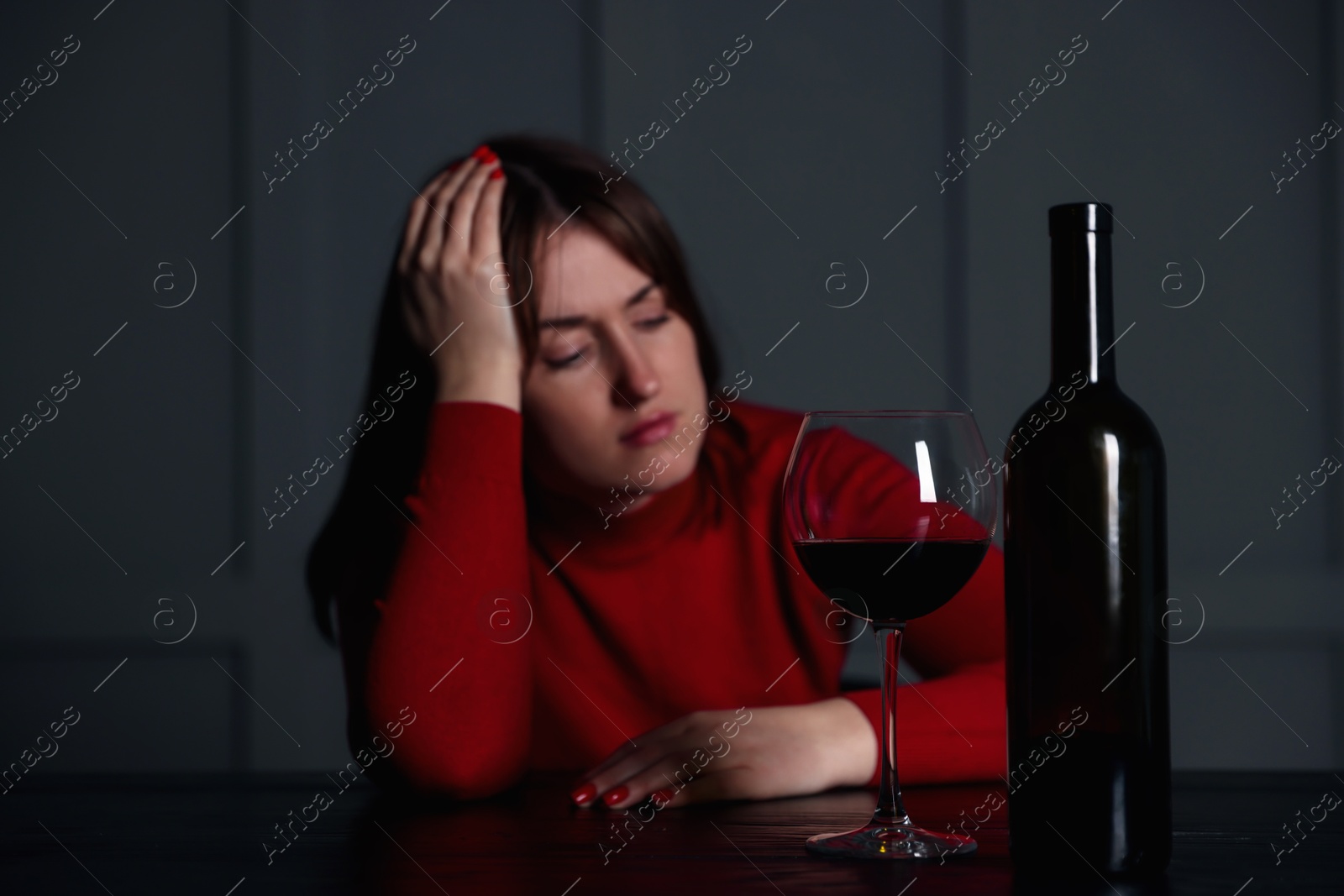 Photo of Alcohol addiction. Woman at wooden table indoors, focus on glass of red wine and bottle