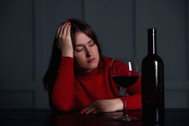 Photo of Alcohol addiction. Woman at wooden table indoors, focus on glass of red wine and bottle