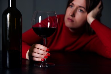 Photo of Alcohol addiction. Woman with glass of red wine and bottle at wooden table indoors, selective focus