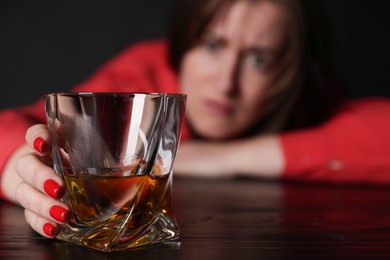 Photo of Alcohol addiction. Miserable woman with whiskey at table in dark, selective focus