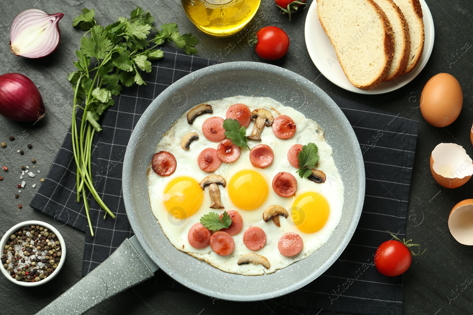 Photo of Tasty fried eggs with cut sausages and mushrooms in frying pan surrounded by ingredients on black table, flat lay