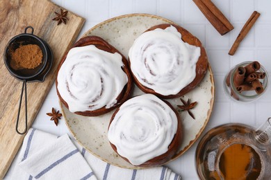 Photo of Tasty cinnamon rolls with cream, spices and tea on white tiled table, flat lay