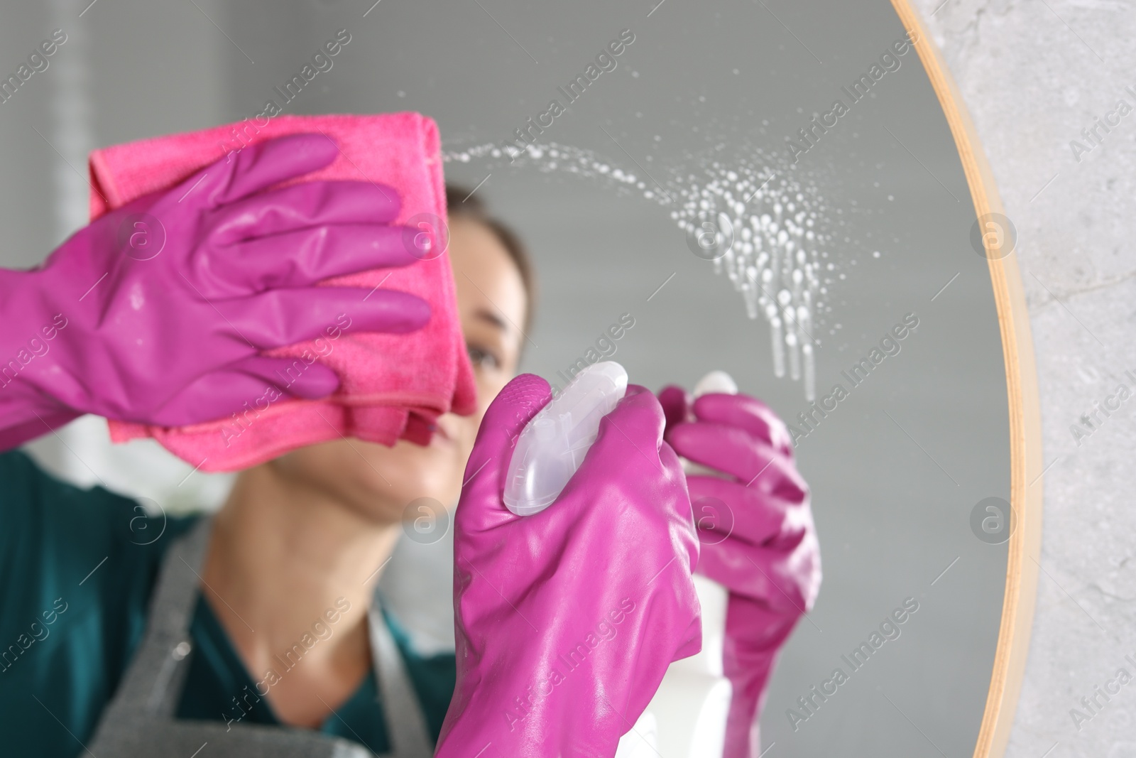 Photo of Woman using cleaning product while wiping mirror with rag indoors