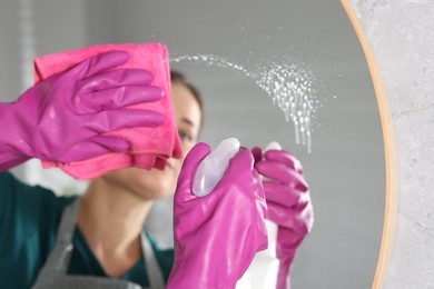 Photo of Woman using cleaning product while wiping mirror with rag indoors