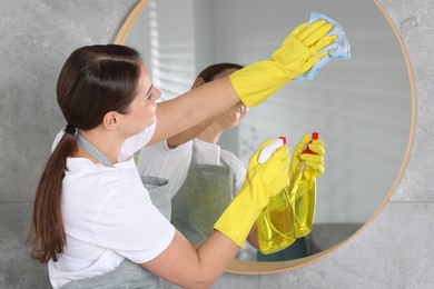 Photo of Woman using cleaning product while wiping mirror with rag indoors