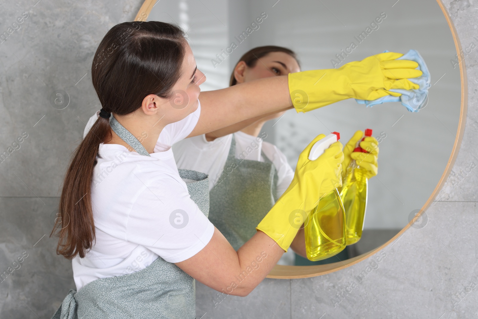 Photo of Woman using cleaning product while wiping mirror with rag indoors