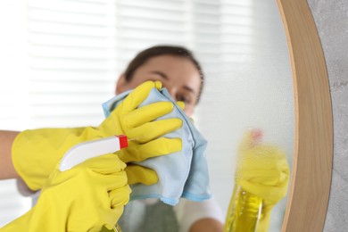 Photo of Woman using cleaning product while wiping mirror with rag indoors