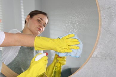 Photo of Woman using cleaning product while wiping mirror with rag indoors