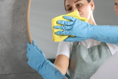 Photo of Woman in gloves cleaning mirror with rag indoors