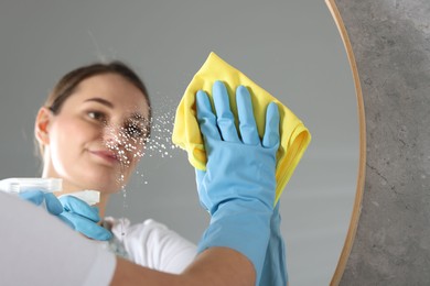 Photo of Woman using cleaning product while wiping mirror with rag indoors