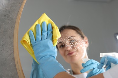 Photo of Woman using cleaning product while wiping mirror with rag indoors