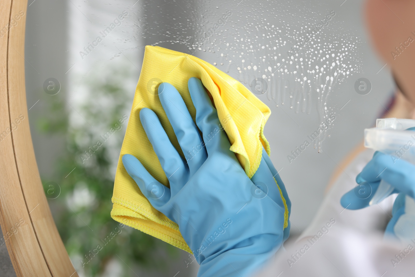 Photo of Woman using cleaning product while wiping mirror with rag indoors, closeup