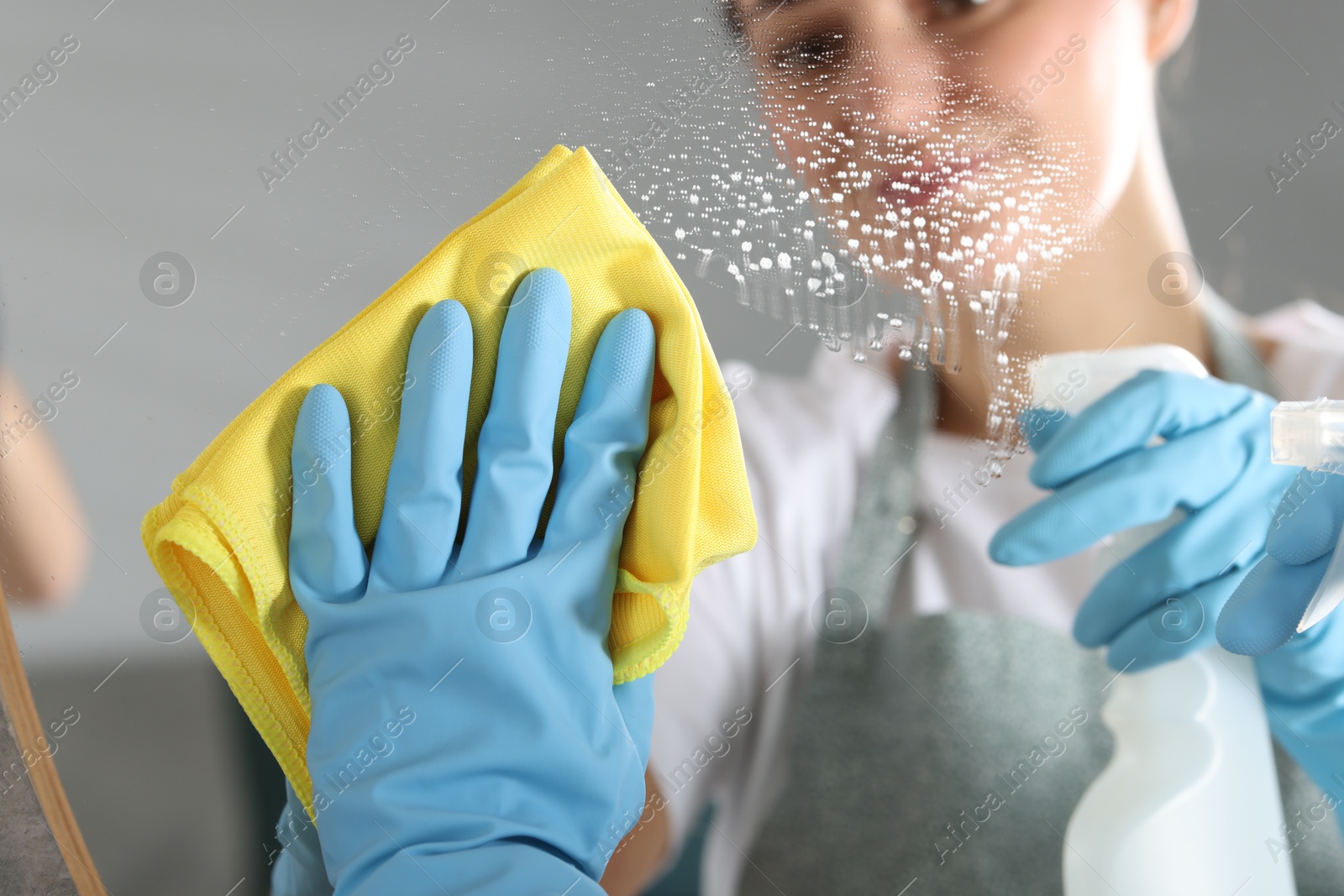 Photo of Woman using cleaning product while wiping mirror with rag indoors, closeup