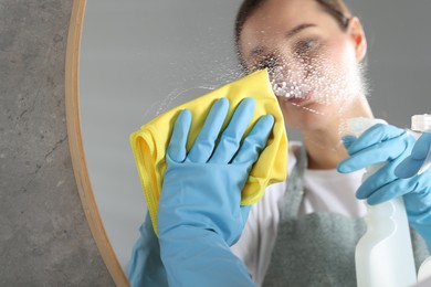 Photo of Woman using cleaning product while wiping mirror with rag indoors