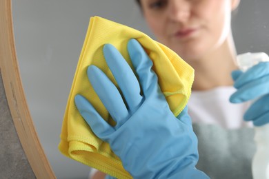 Photo of Woman using cleaning product while wiping mirror with rag indoors, closeup