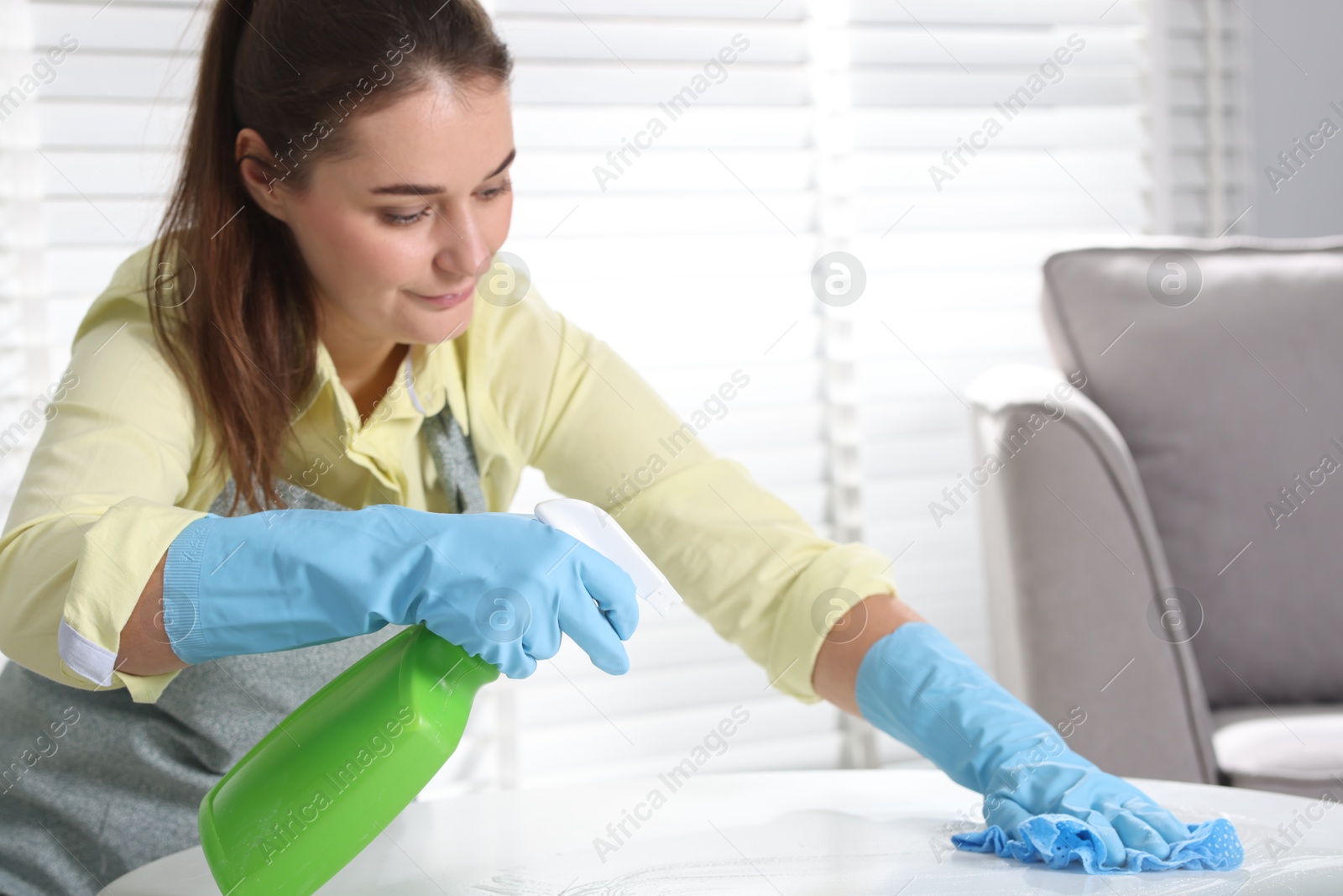 Photo of Woman using cleaning product while wiping table with rag indoors