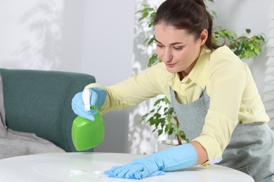 Photo of Woman using cleaning product while wiping table with rag indoors