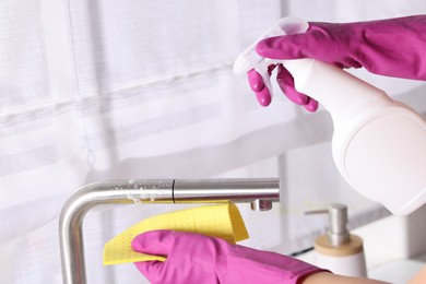 Photo of Woman using cleaning product while wiping faucet of kitchen sink with rag indoors, closeup