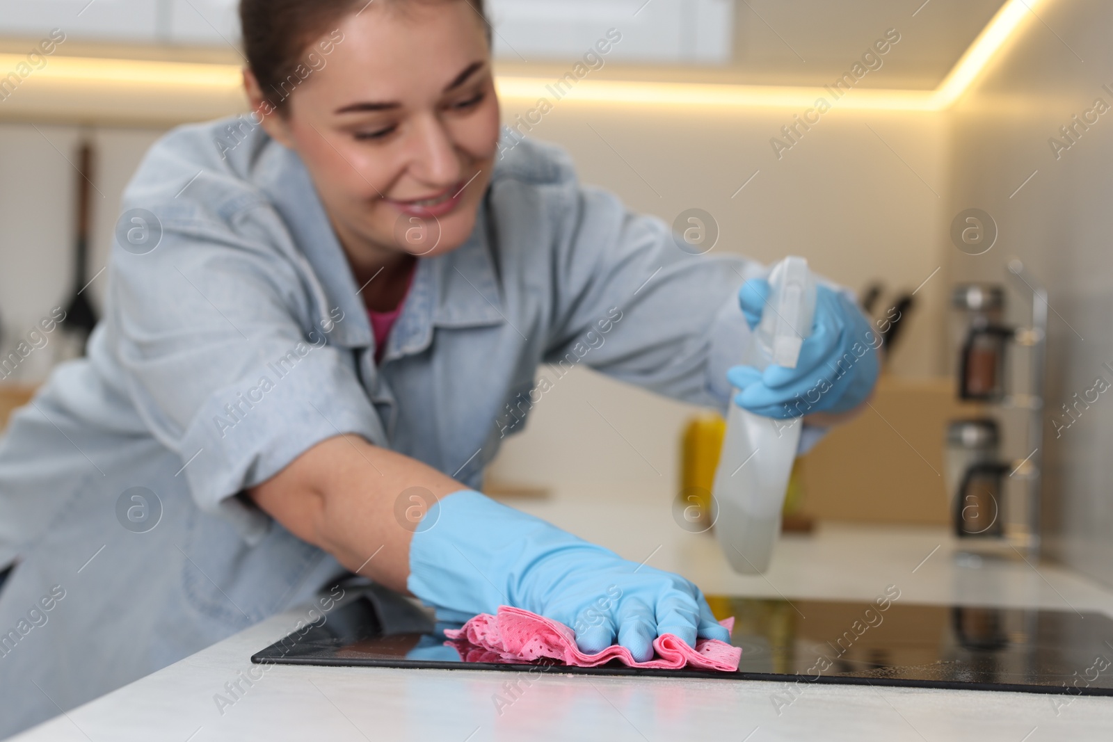 Photo of Woman using cleaning product while wiping induction cooktop with rag in kitchen, selective focus. Space for text