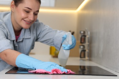 Photo of Woman using cleaning product while wiping induction cooktop with rag in kitchen, selective focus. Space for text