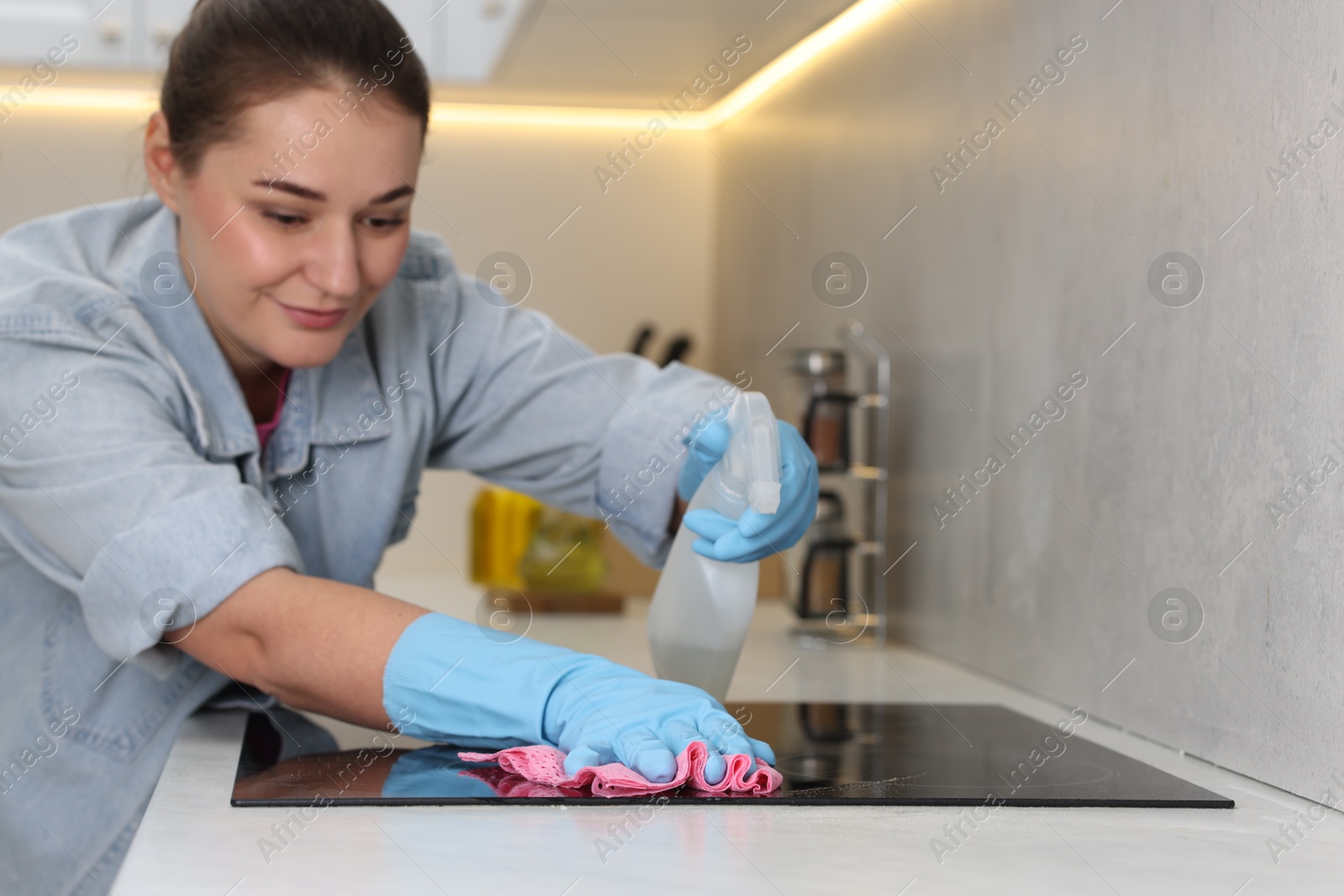 Photo of Woman using cleaning product while wiping induction cooktop with rag in kitchen, selective focus. Space for text