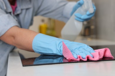 Photo of Woman using cleaning product while wiping induction cooktop with rag in kitchen, closeup