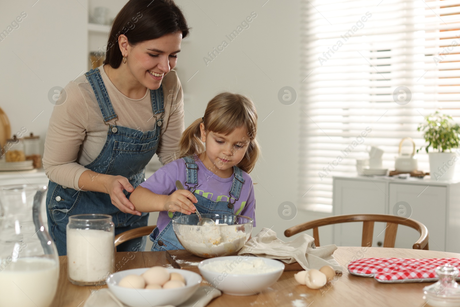 Photo of Little girl helping her mom making dough at table in kitchen, space for text