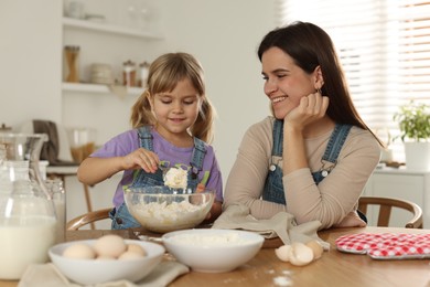 Photo of Little girl helping her mom making dough at table in kitchen