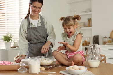 Little girl helping her mom making dough at table in kitchen