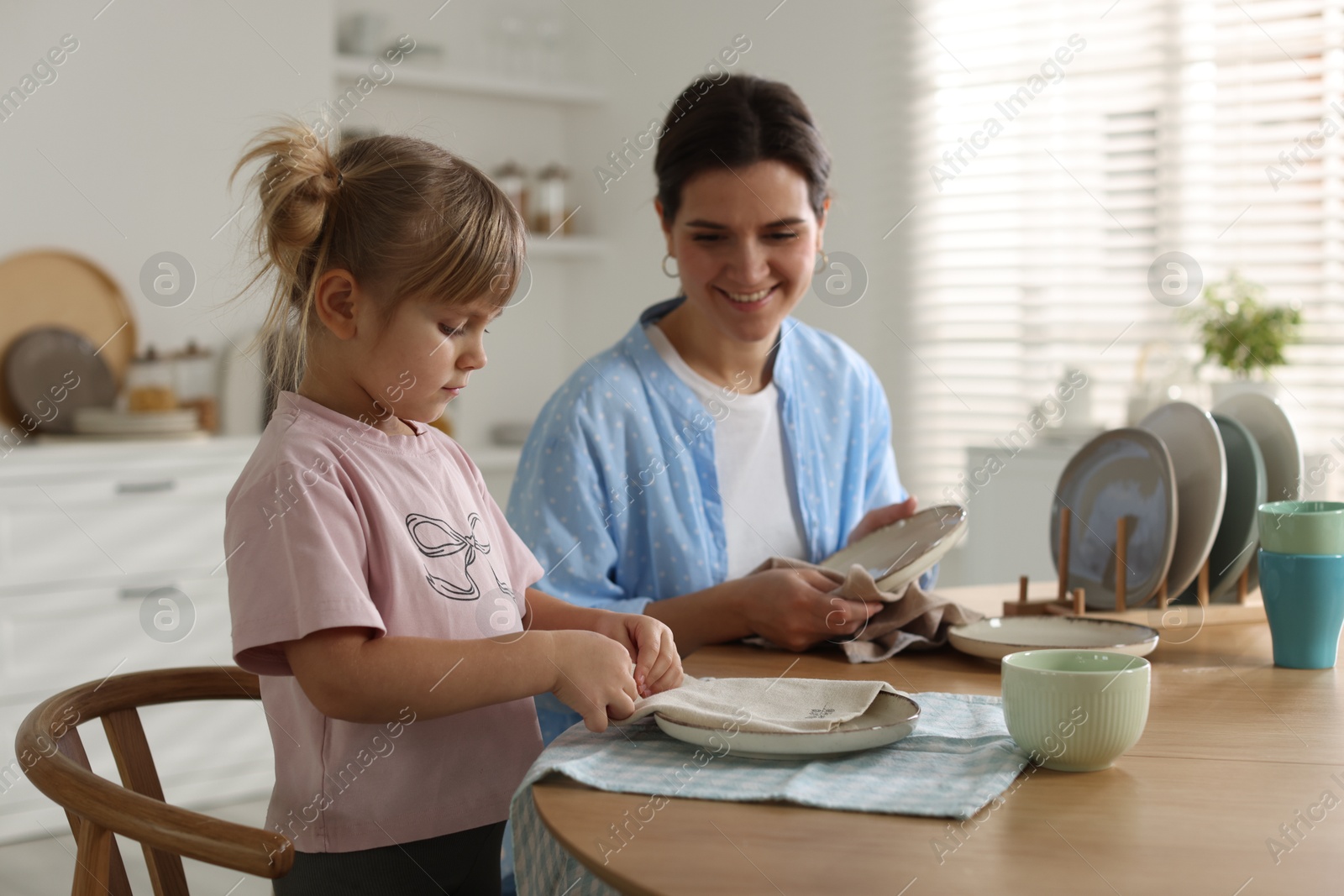 Photo of Little girl helping her mom wiping plates at table in kitchen