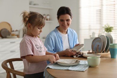 Little girl helping her mom wiping plates at table in kitchen