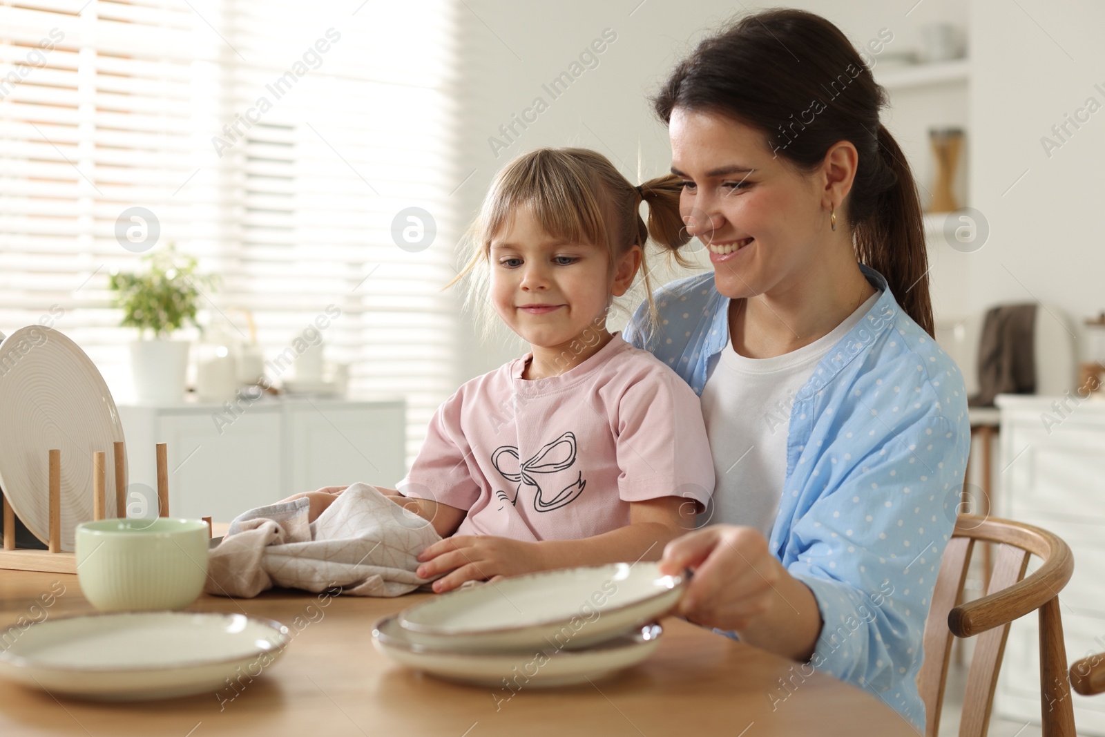 Photo of Little girl helping her mom wiping plates at table in kitchen