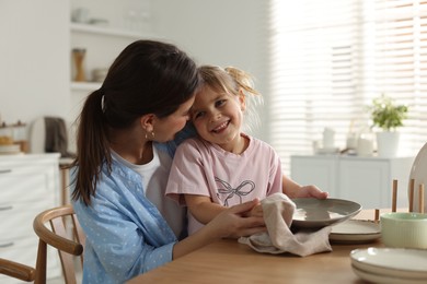 Photo of Little girl helping her mom wiping plates at table in kitchen