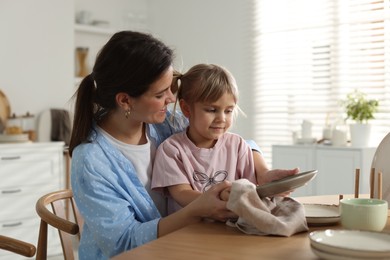 Photo of Little girl helping her mom wiping plates at table in kitchen