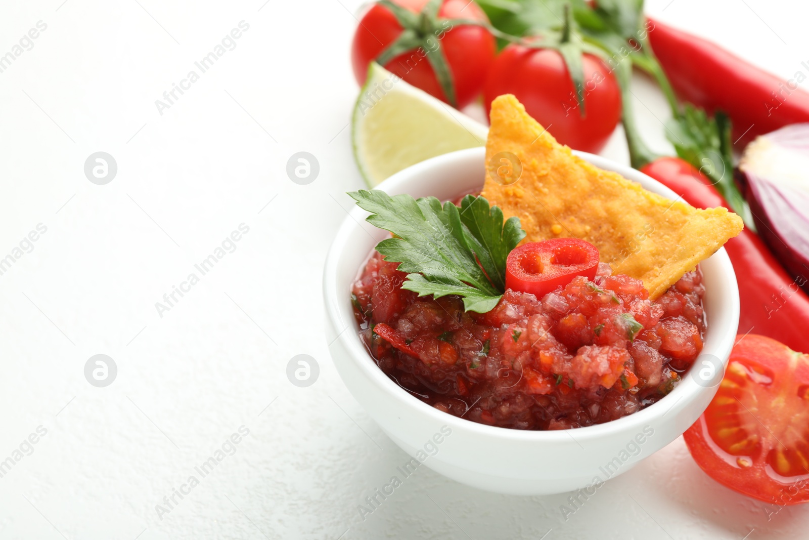 Photo of Spicy salsa in bowl, nacho chip and ingredients on white table, closeup. Space for text