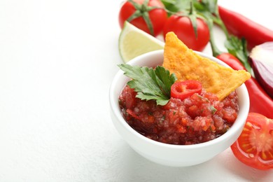 Spicy salsa in bowl, nacho chip and ingredients on white table, closeup. Space for text