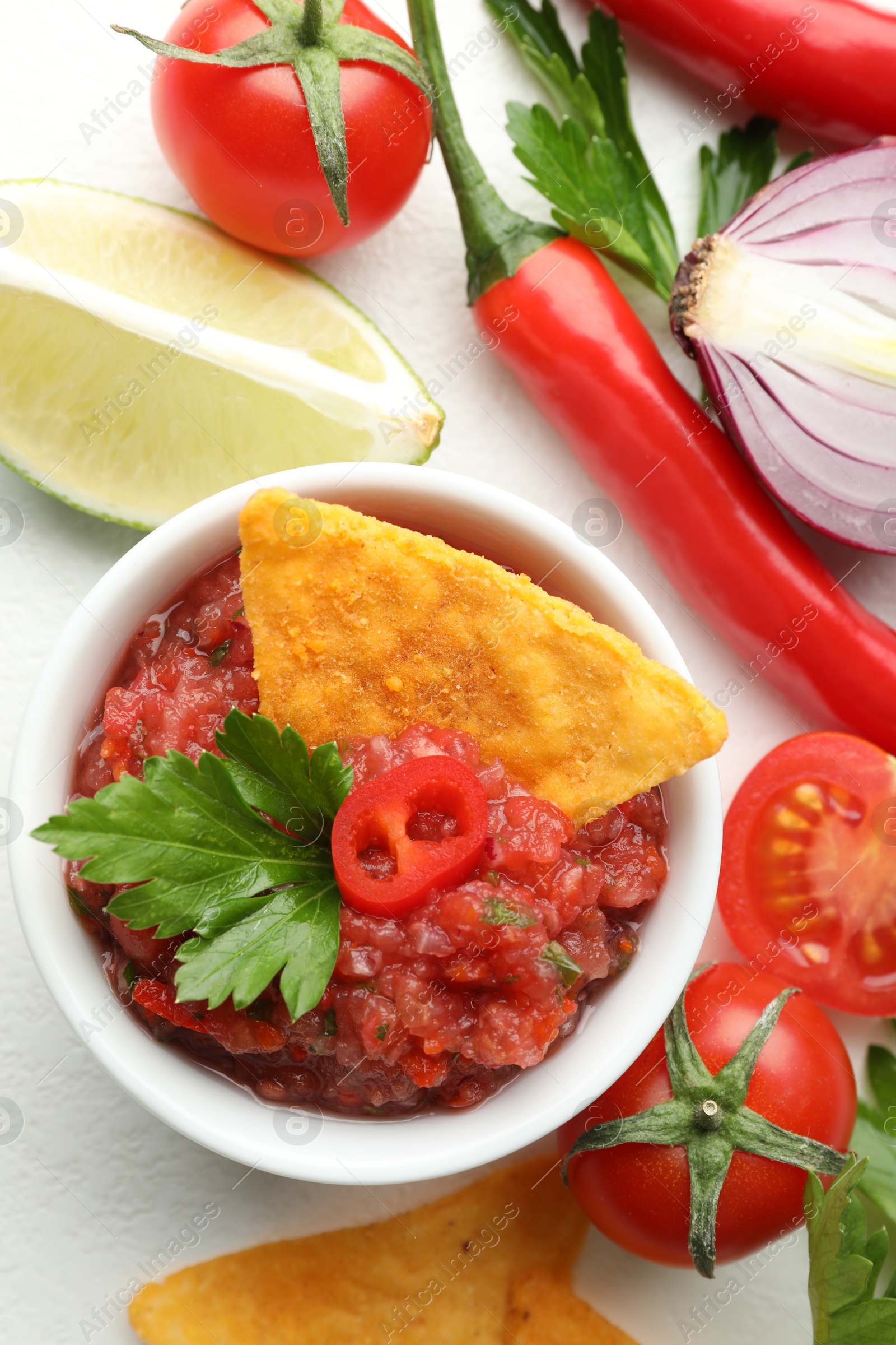 Photo of Spicy salsa in bowl, nachos and ingredients on white table, flat lay