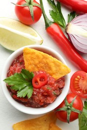 Spicy salsa in bowl, nachos and ingredients on white table, flat lay