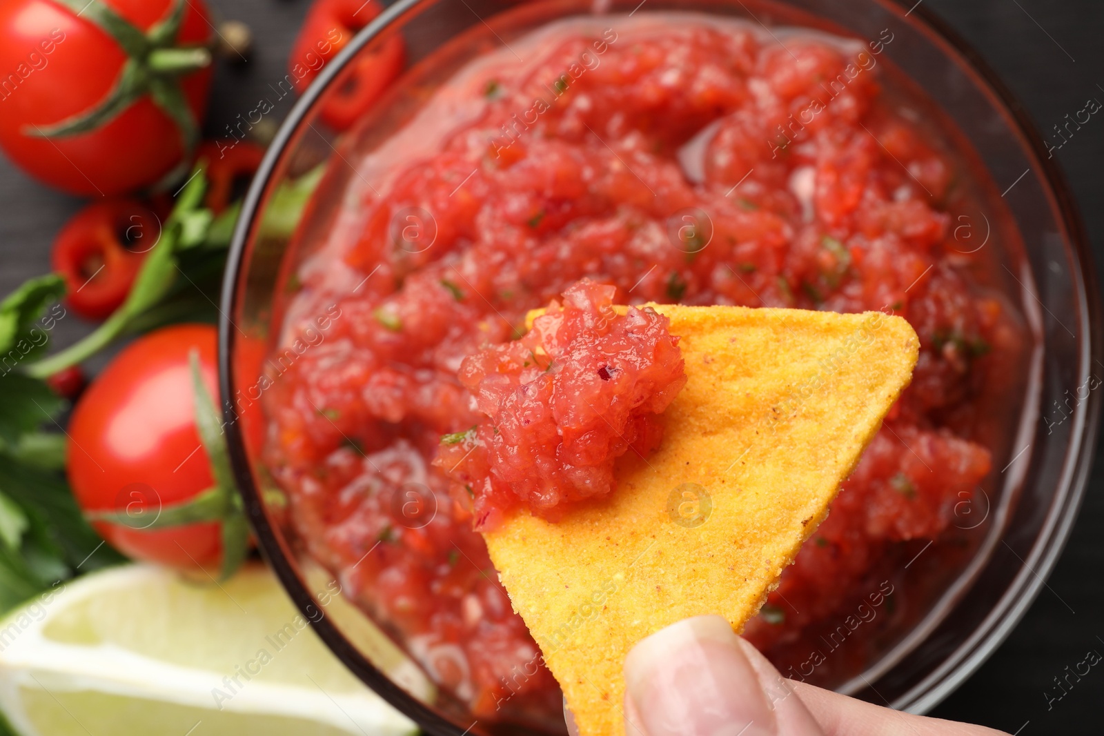 Photo of Woman dipping nacho chip into spicy salsa at table, top view