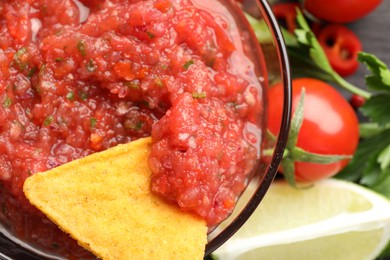 Spicy salsa in bowl, nacho chip and ingredients on table, top view