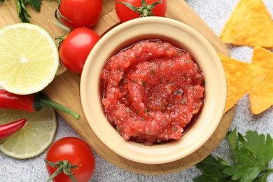 Spicy salsa in bowl, nachos and ingredients on light grey table, flat lay