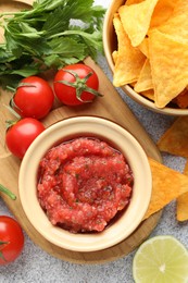 Spicy salsa in bowl, nachos and ingredients on light grey table, flat lay