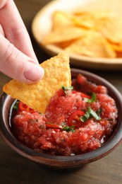 Photo of Woman dipping nacho chip into spicy salsa at table, closeup