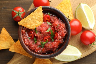 Spicy salsa in bowl, nachos, tomatoes and lime on wooden table, flat lay