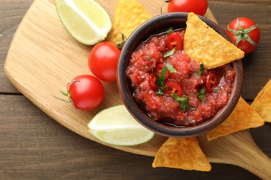 Spicy salsa in bowl, nachos, tomatoes and lime on wooden table, flat lay