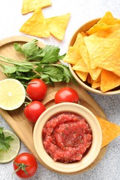 Spicy salsa in bowl, nachos and ingredients on light grey table, flat lay