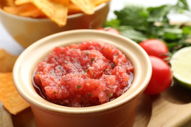 Spicy salsa in bowl, nachos and ingredients on table, closeup